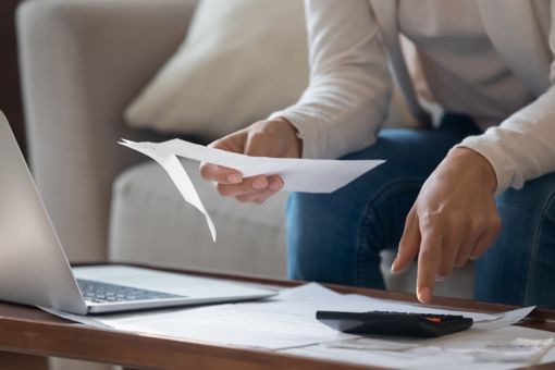A hand is typing something on a calculator and holding a piece of paper in front of a laptop.